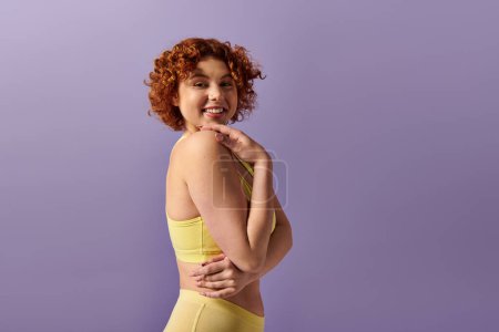 Curvy young redhead in yellow lingerie striking a pose against a purple backdrop.