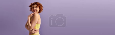 A young, curvy redhead woman poses beautifully in a yellow underwear against a vibrant purple backdrop.