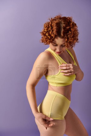 A young, curvy redhead woman in a striking yellow bikini strikes a confident pose against a vibrant purple background.