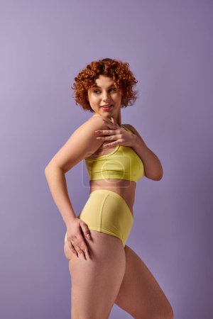Curvy redhead in yellow underwear poses against a vivid purple backdrop.