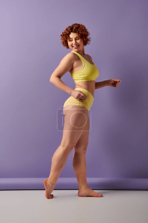 A young, curvy redhead woman in a yellow bikini striking a pose against a bold purple wall.
