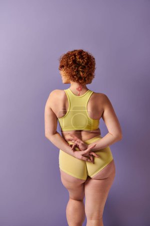Young redhead woman flaunts curves in yellow bikini against vibrant purple backdrop.