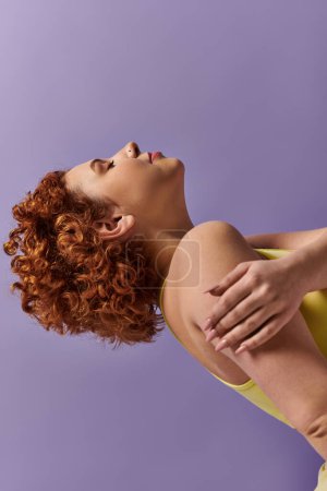 A curvy young redhead woman, dressed in a yellow shirt, performs a back stretch against a purple background.