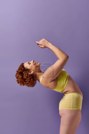 Curvy redhead woman in yellow bikini posing gracefully against a vibrant purple wall.