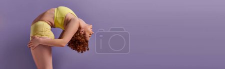 A young, curvy redhead woman in yellow underwear gracefully executes a yoga pose on a vibrant purple background.
