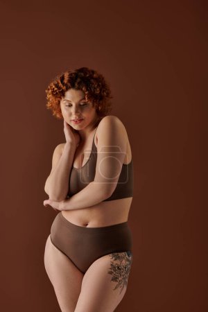 Foto de A curvy redhead woman strikes a pose in a brown bikini against a matching background. - Imagen libre de derechos