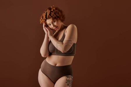 Young, curvy redhead woman exudes confidence in brown bikini on brown background.