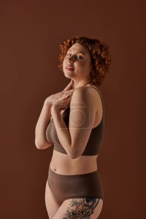 A young, curvy redhead woman confidently poses in a brown bikini.