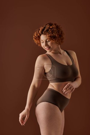 A curvaceous redhead woman is confidently posing in a brown bikini.