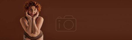 Téléchargez les photos : A young, curvy redhead woman exudes confidence in a bikini, striking a pose on a plain brown background. - en image libre de droit