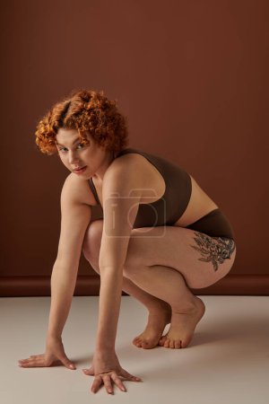 A crouching young, curvy woman with fiery red hair on a brown background.