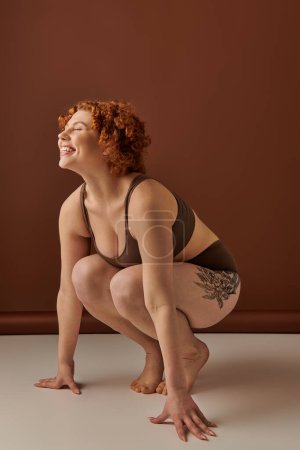 Young, curvy redhead woman crouches gracefully in brown underwear on a textured earth-toned background.