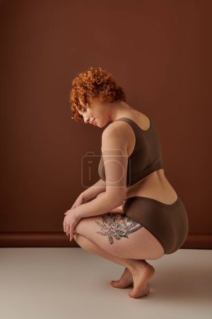 A young, curvy redhead woman crouches in brown underwear, exuding a mix of strength and vulnerability.