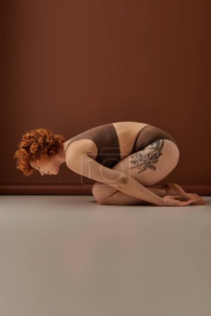 A curvy redhead crouches on the floor displaying her vibrant tattoos.