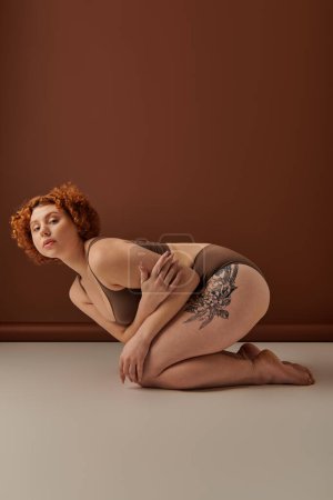 A young curvy redhead woman in underwear crouches on a brown background.