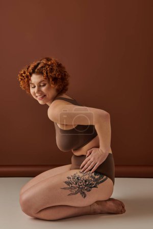 Curvy redhead woman sitting on floor with thigh tattoo.
