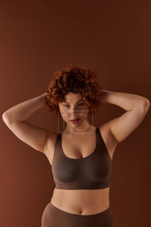 A young curvy redhead woman strikes a pose in a brown sports bra against a neutral background.