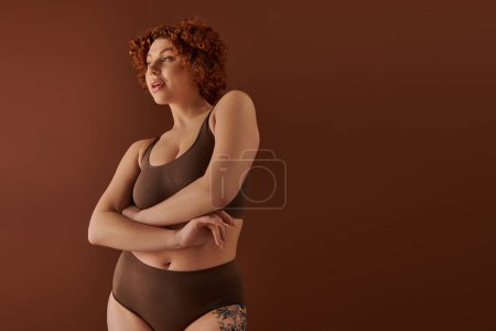 A young, curvy redhead woman poses in a stylish brown bikini against a complementary background, exuding confidence and allure.