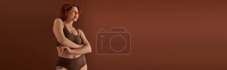 Foto de A young, curvy redhead woman in a bikini exudes confidence in front of a brown background. - Imagen libre de derechos