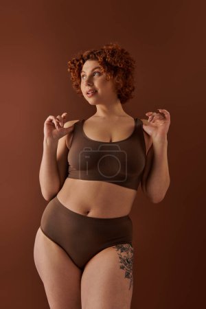 Téléchargez les photos : A young and curvy redhead woman in a brown bikini poses gracefully on a warm-toned brown background. - en image libre de droit