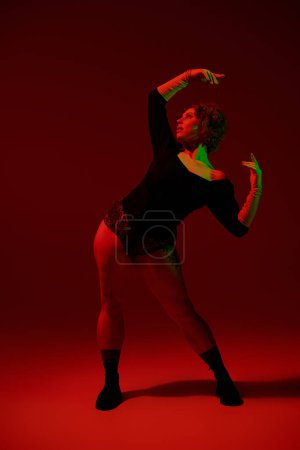 A young curvy redhead woman in a bodysuit dances gracefully in front of a vivid red background.