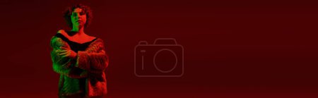 Téléchargez les photos : A young, curvy redhead woman in a fur coat stands boldly in front of a rich red background. - en image libre de droit