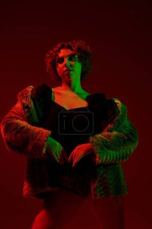 Photo for A young curvy redhead woman in a fur coat striking a pose in front of a vibrant red light. - Royalty Free Image