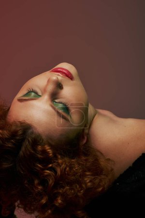 A young, curvy redhead woman in a bodysuit is laying down, exuding an ethereal aura under soft red lighting.