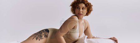 Photo for A young, curvy redhead woman in lingerie reclines on a bed with a tattoo, against a grey background. - Royalty Free Image
