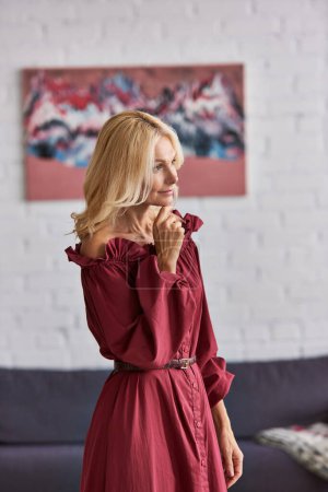 Foto de Una mujer madura en un vestido rojo llamativo se levanta con gracia en una acogedora sala de estar. - Imagen libre de derechos