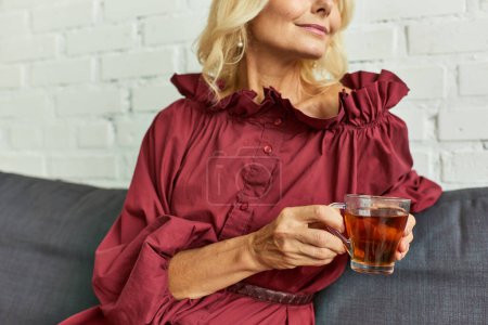 Femme mûre en robe élégante profitant d'un moment de détente avec un verre de thé sur un canapé.