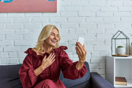 Mature woman in stylish dress, holding cell phone on couch.
