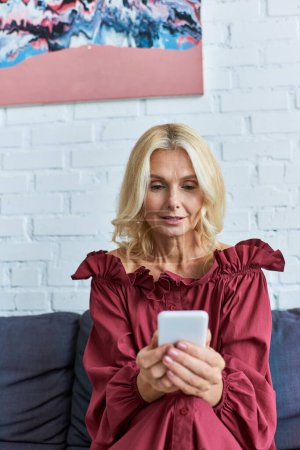 Photo for A mature woman in a stylish dress absorbed in her cell phone on a couch. - Royalty Free Image