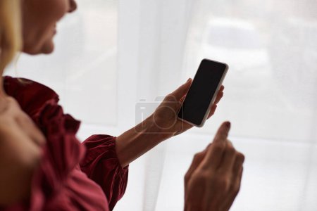 Téléchargez les photos : Femme mûre dans une chemise rouge tient élégamment un téléphone portable à la maison. - en image libre de droit