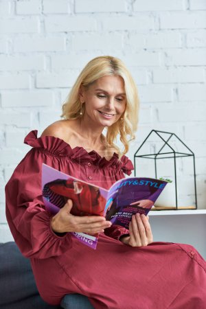Téléchargez les photos : Femme élégante en tenue élégante absorbée dans un magazine, assise sur un canapé confortable. - en image libre de droit