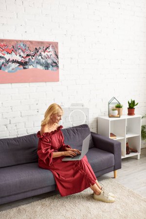 Mature woman in stylish attire sits on couch, engrossed in laptop usage.