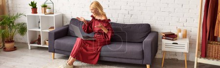 Foto de Una mujer madura en un vestido elegante sentado en un sofá con ordenador portátil. - Imagen libre de derechos