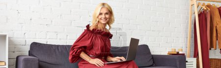 Téléchargez les photos : Femme mûre en robe élégante en utilisant un ordinateur portable sur le canapé. - en image libre de droit