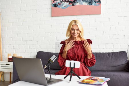 Foto de Mujer madura en vestido rojo chic creando un podcast sobre la belleza femenina delante de un ordenador portátil. - Imagen libre de derechos
