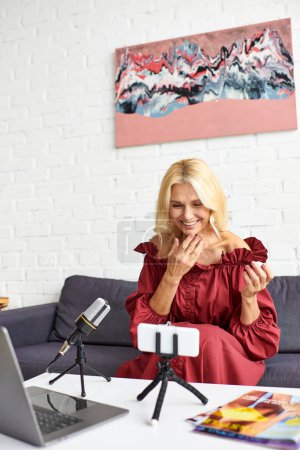 Eine reife elegante Frau in einem roten Kleid sitzt an einem Tisch vor einem Laptop und nimmt einen Podcast über weibliche Schönheit auf.