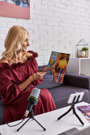 Foto de Una mujer madura y elegante con un vestido rojo se sienta en un sofá, absorto en la lectura de una revista y la creación de podcast. - Imagen libre de derechos