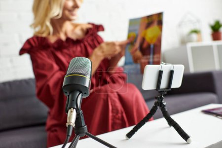 Photo for A mature woman in a chic red dress sits on a couch, speaking into a microphone. - Royalty Free Image