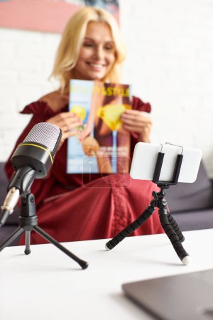 A mature elegant woman in a red chic dress sitting in front of a microphone, holding a magazine.