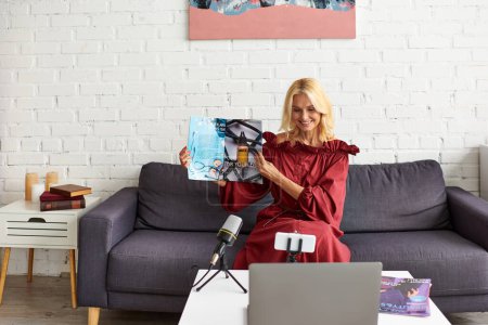 Mature elegant woman in red chic dress records a podcast on female beauty seated in front of a laptop on a couch.