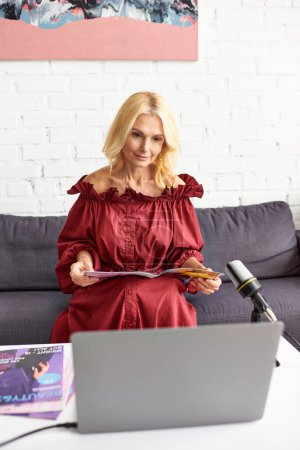Reife Frau im roten Kleid zeichnet Podcast über weibliche Schönheit mit Laptop auf.