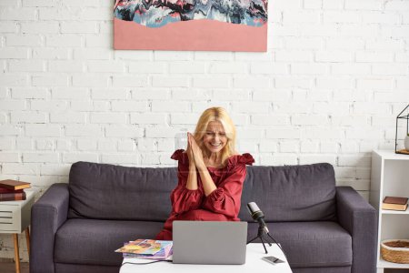 Mujer madura elegante en vestido rojo chic sentado en un sofá delante de un ordenador portátil, creando un podcast sobre la belleza femenina.