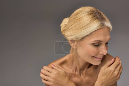 Fröhliche reife Frau mit blonden Haaren posiert vor grauem Hintergrund.