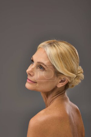 Reife Frau mit blonden Haaren im Zopf vor grauem Hintergrund.