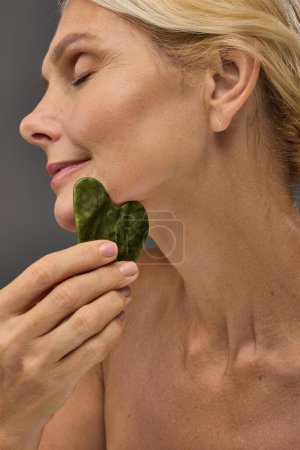 Photo for Attractive mature woman using gua sha on a gray backdrop. - Royalty Free Image
