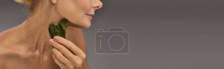 Téléchargez les photos : Séduisante femme mature en utilisant gua sha sur une toile de fond grise. - en image libre de droit